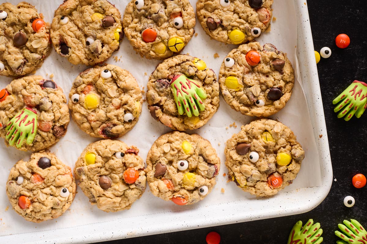 A baking sheet with baked monster cookies on a dark background with decorated with zombie hands, candy eyes and M&Ms