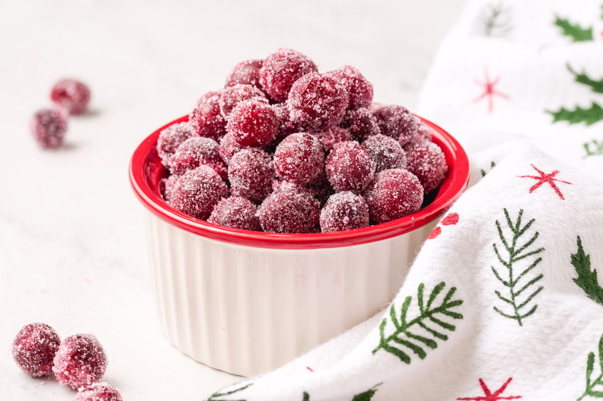 Bowl of sugared cranberries with a red rim with a Christmas themed towel wrapped around it on a white marble background with a few cranberries on the table