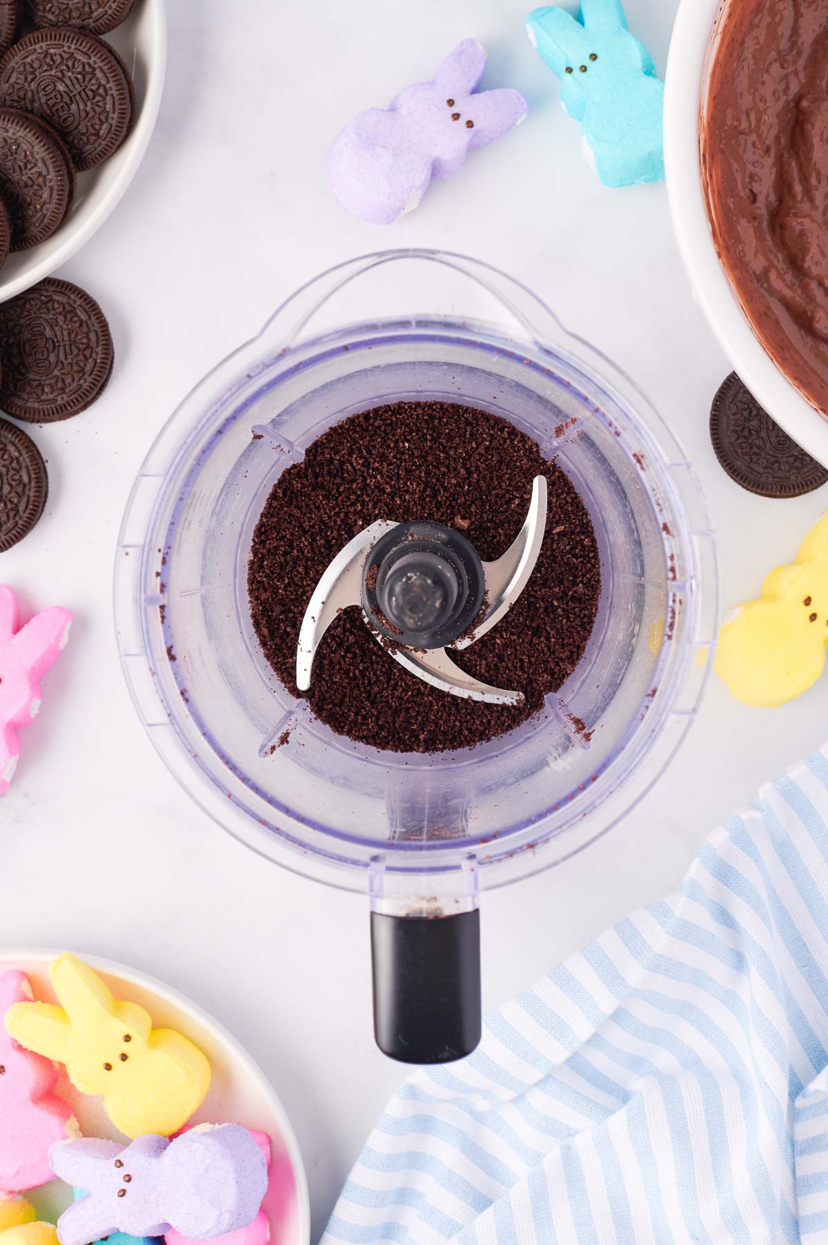 Oreos pulsed into crumbs in a food processor on a white background surrounded by Oreo cookies and bunny  Peeps marshmallows