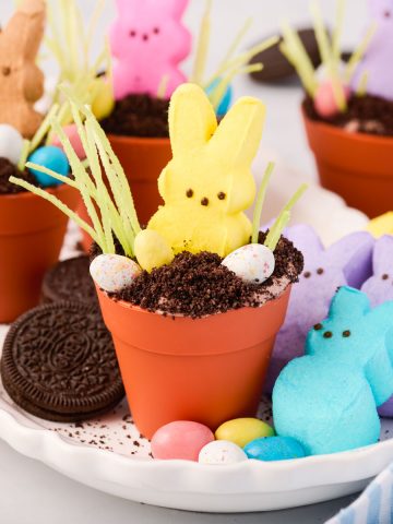Easter Dirt Cups decorated with a yellow Peeps bunny, green candy grass and chocolate eggs on a serving platter surrounded by Oreos, bunny Peeps, and Whopper candy eggs