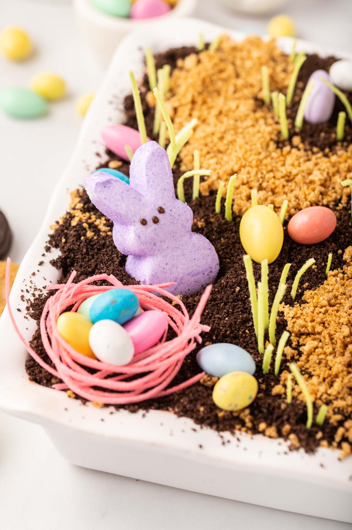 close up of a nest created with edible candy grass with chocolate egg candies inside and a purple Peeps marshmallow bunny sticking out of dirt cake