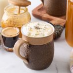 A honey lavender latte in a brown mug with a shot of espresso and pot of honey sitting next to it