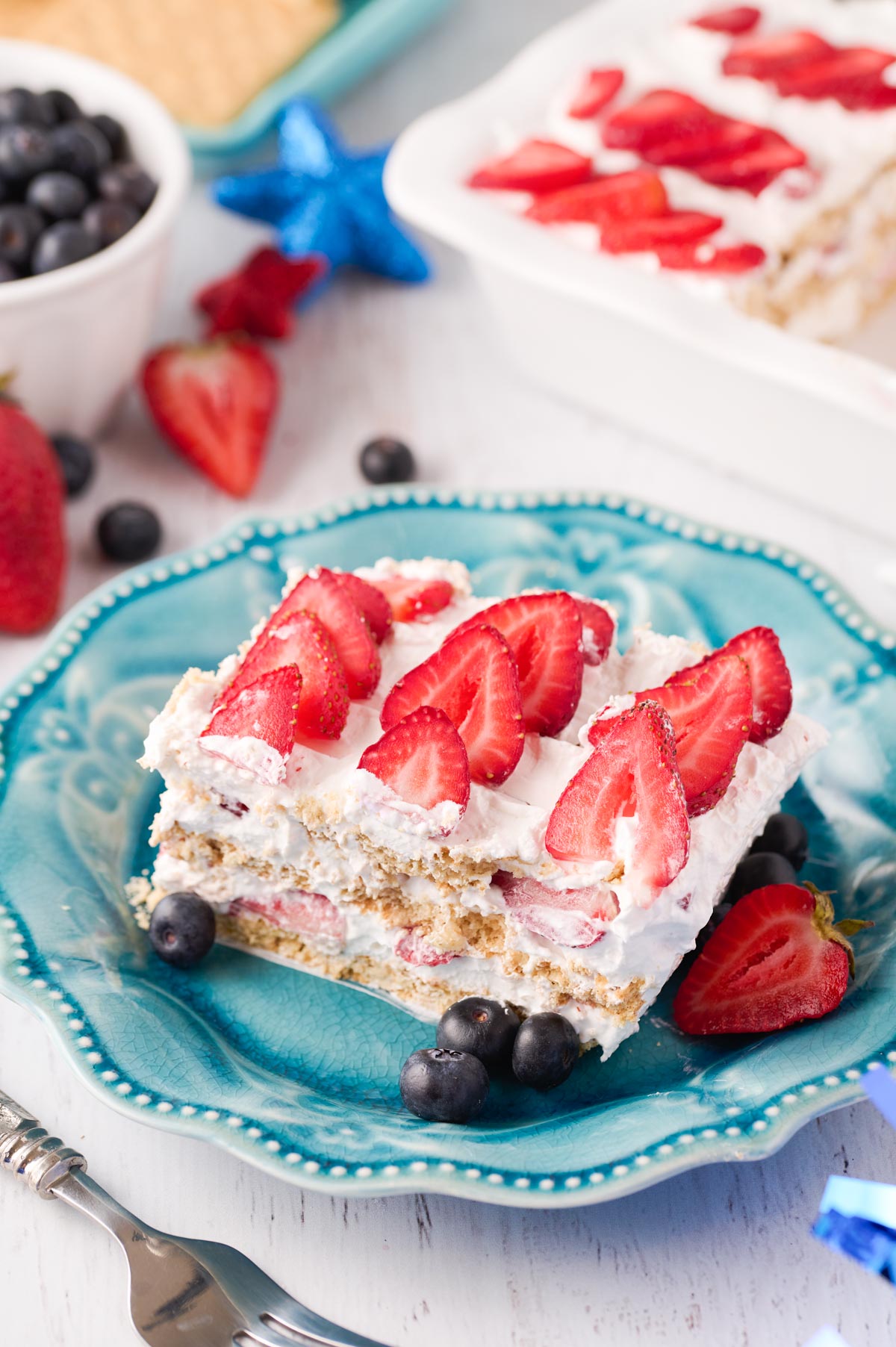 Slice of strawberry icebox cake sits on a blue plate with fresh blueberries and strawberries