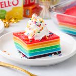 A slice of rainbow jello cake sits on a white plate topped with cool whip and rainbow sprinkles