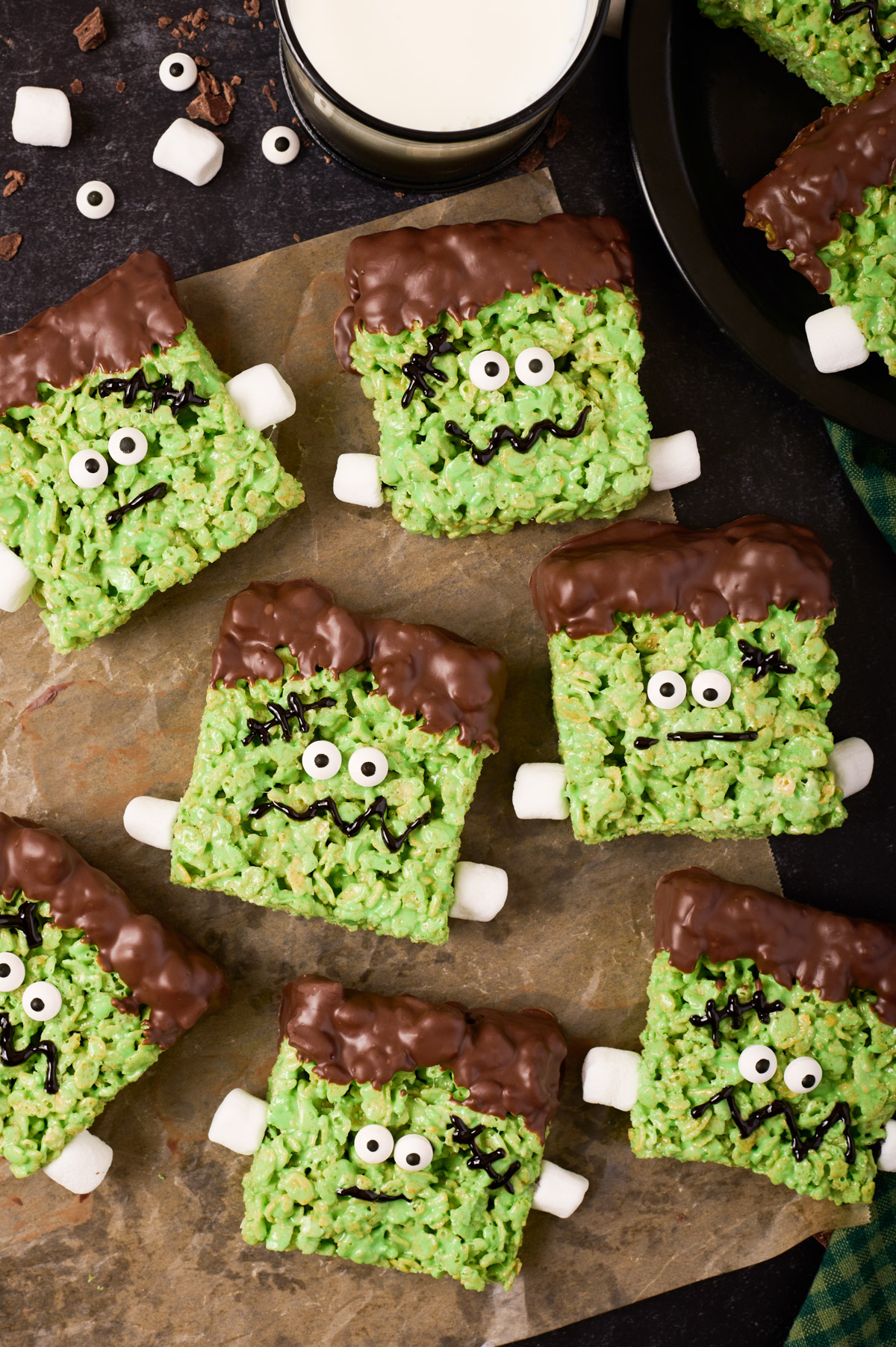 Frankenstein rice krispie treats sit on a piece of wax paper surrounded by more rice krispie treats, candy eyes, chocolate pieces, a glass of milk and green linen on a black background