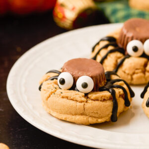 Three spider peanut butter cookies sit on a small white black on a dark background surrounded by more cookies and ingredients