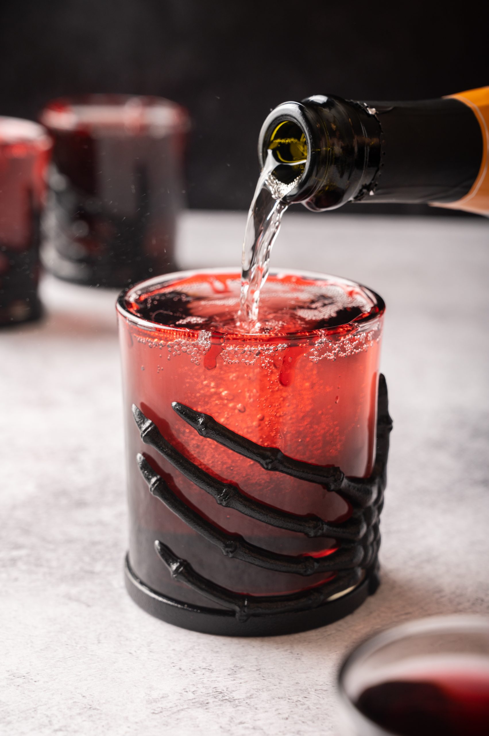 Topping a Vampire's Kiss Cocktail with Prosecco in a glass with a black skeleton hand wrapped around it