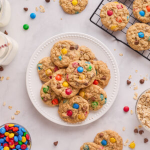 A plate of monster cookies is surrounded by ingredients to make the cookies, a wire rack with cookies cooling on it and a glass of milk
