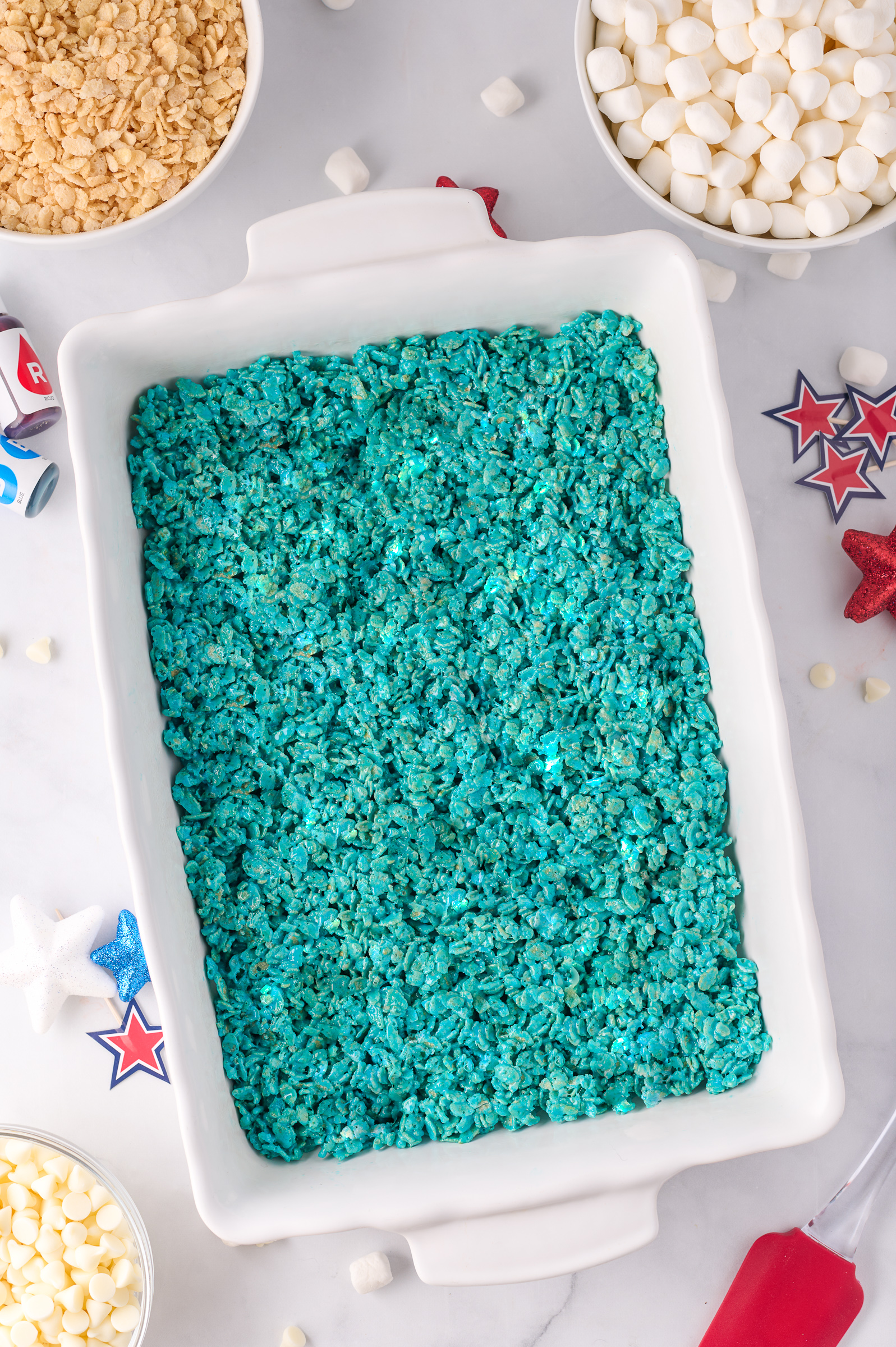 Marshmallow rice krispie mix dyed blue pressed into a white 9 x 13 baking dish with the recipe ingredients surrounding it.