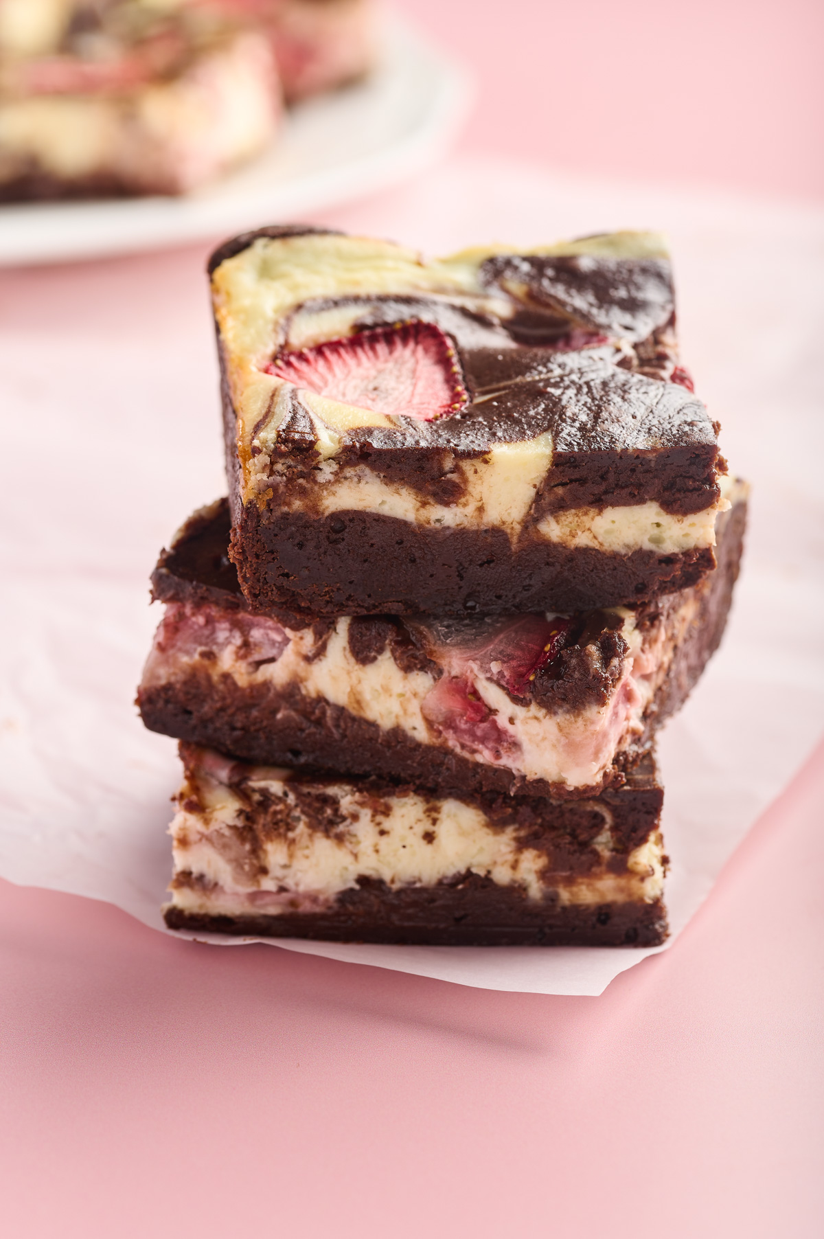 A stack of strawberry cheesecake brownies stands on top of parchment paper with a platter of brownies in the background.