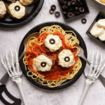 Skeleton hands hold a spoon in one hand and a fork in another on each side of a plate filled with spaghetti, red sauce and halloween eyeball meatballs on top.